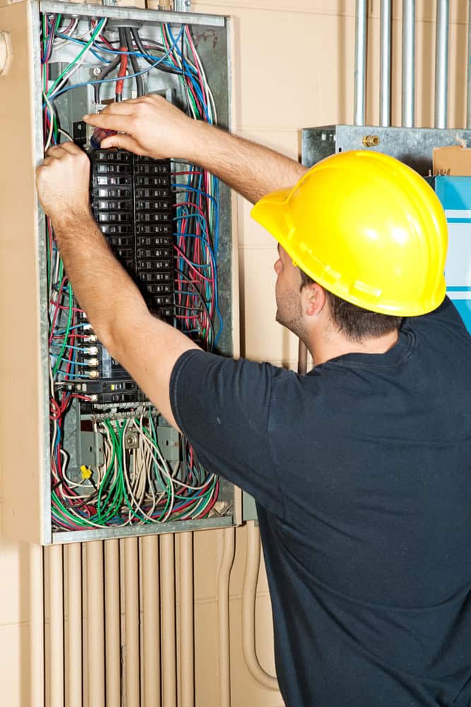 A person wearing a yellow hard hat works on an open electrical panel filled with wires and circuit breakers. A person wearing a yellow hard hat works on an open electrical panel filled with wires and circuit breakers.