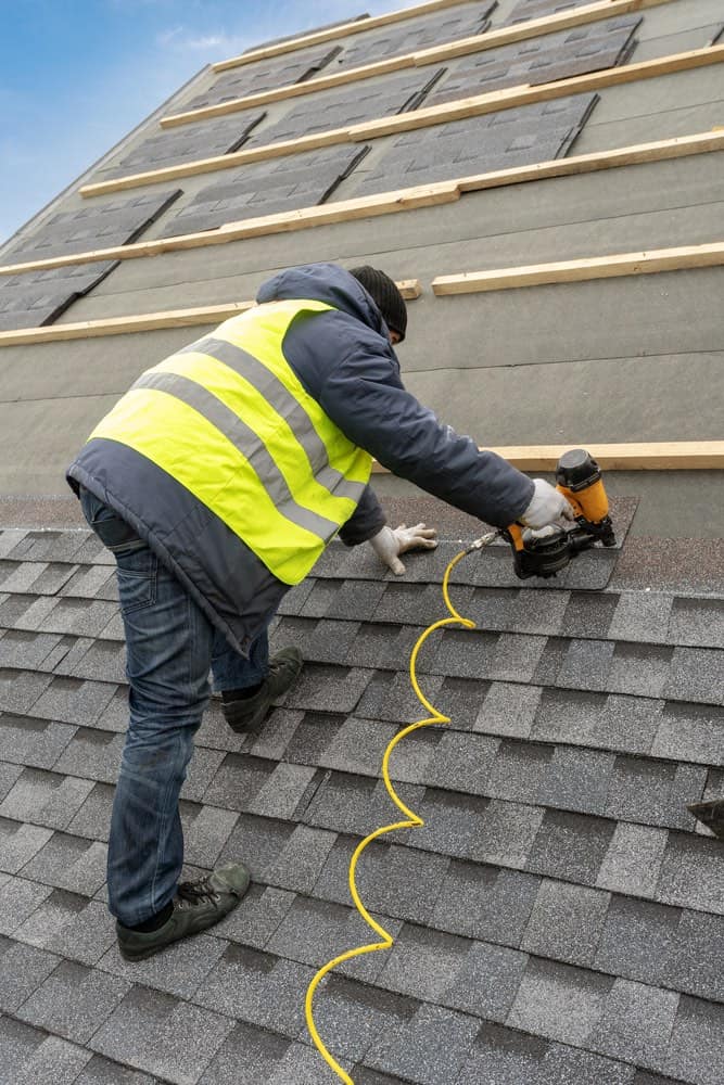 A worker in a high-visibility vest uses a nail gun to install shingles on a residential roof.
